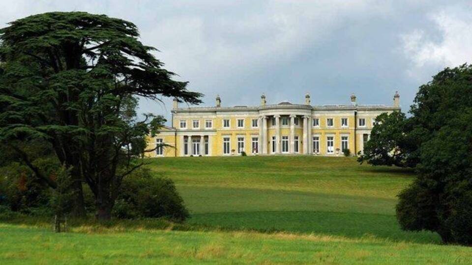 A large, classical building with a symmetrical facade, surrounded by lush greenery and a well-maintained lawn.
