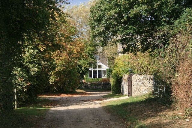 A rural pathway leading to a stone wall and a house surrounded by trees.