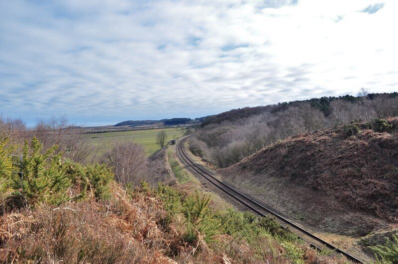 A railway track curving through a hilly landscape with sparse vegetation and a cloudy sky.