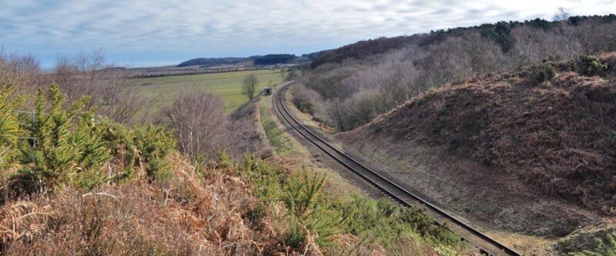 A railway track curving through a hilly landscape with sparse vegetation and a cloudy sky.