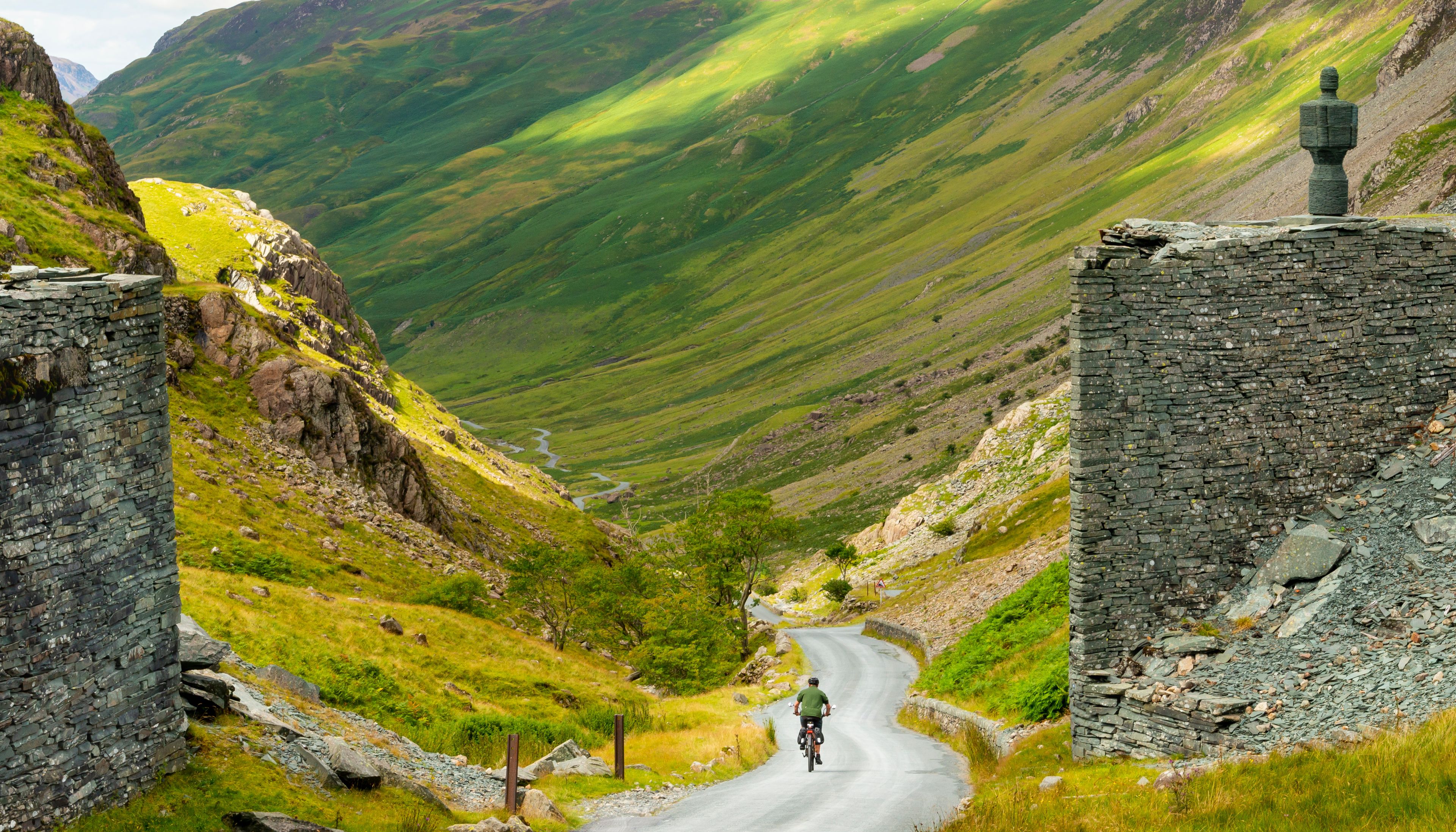 A cyclist on a narrow road flanked by stone ruins and a mountainous landscape.