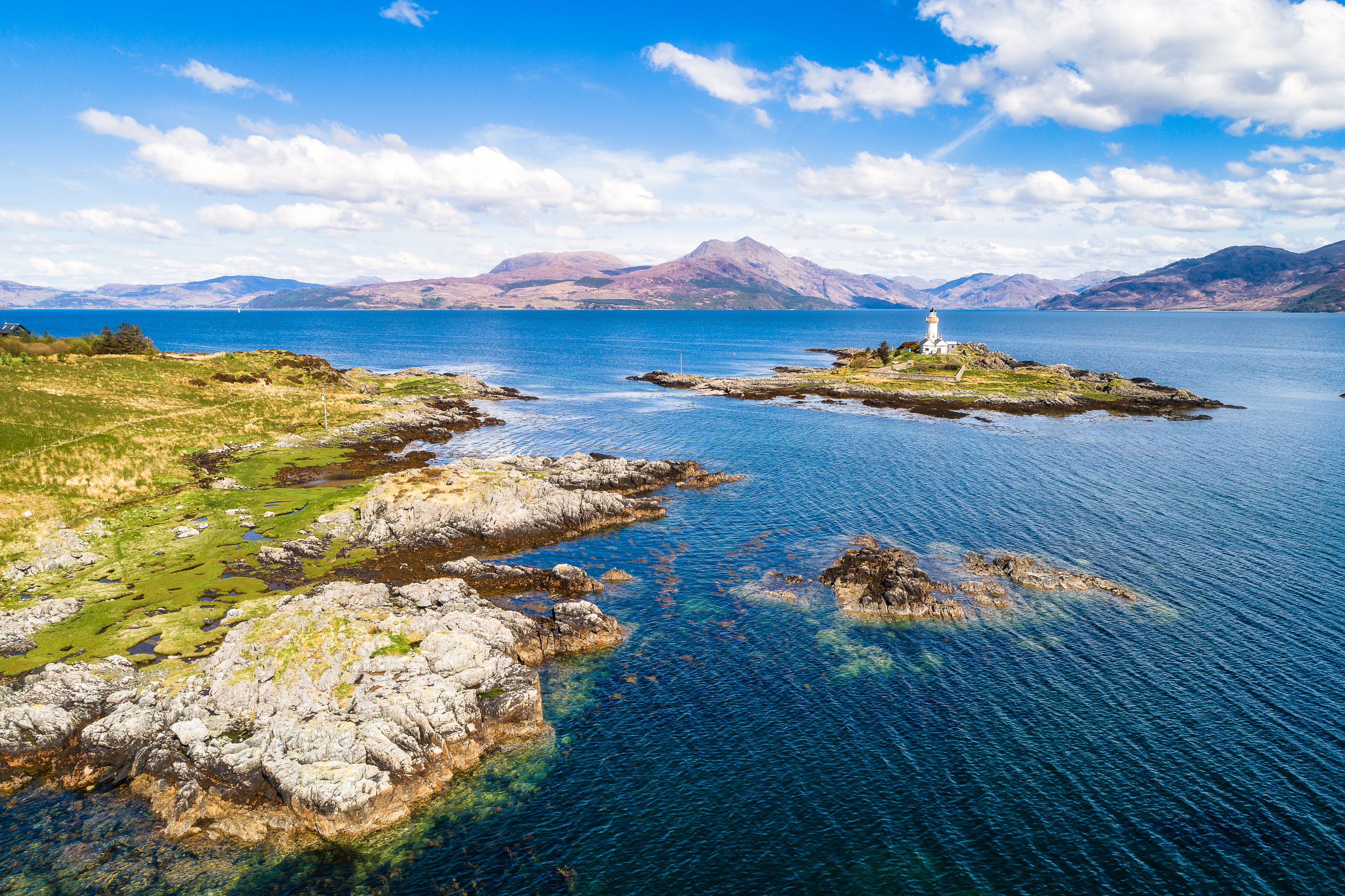 A coastal landscape with a lighthouse on a small island, surrounded by rocky shores and clear blue waters.