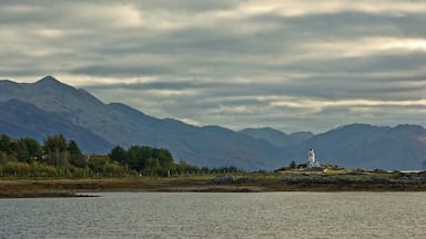 A lighthouse on a rocky outcrop with mountains in the background and a body of water in the foreground.