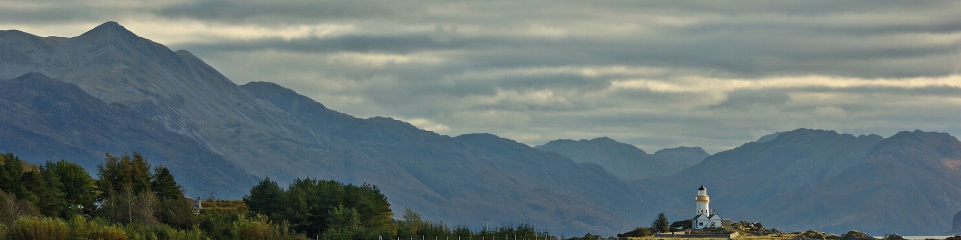 A lighthouse on a rocky outcrop with mountains in the background and a body of water in the foreground.