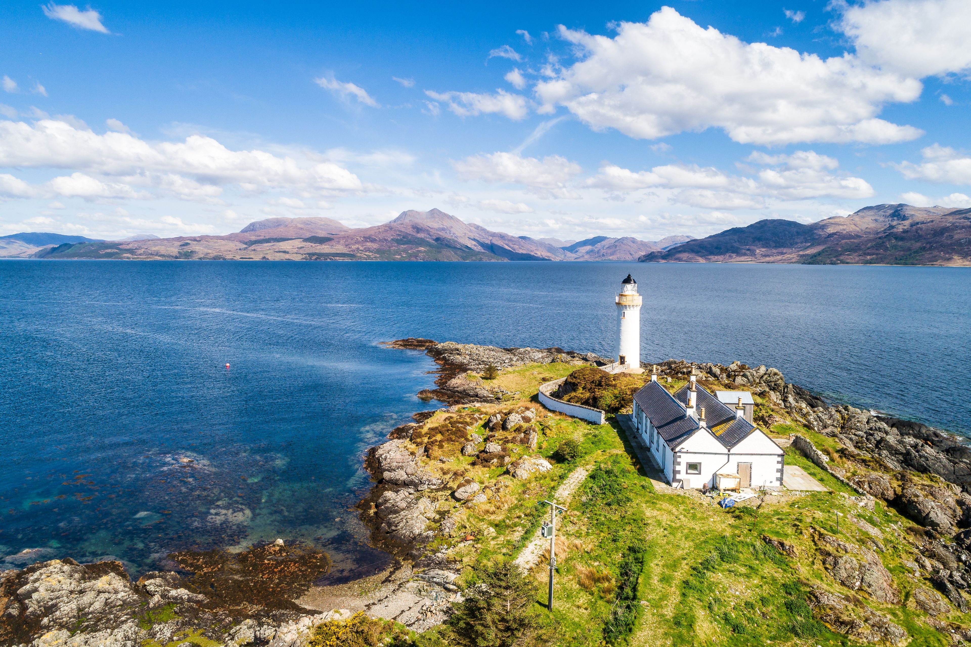 A lighthouse on a rocky coastline with a clear blue sky and mountains in the background.