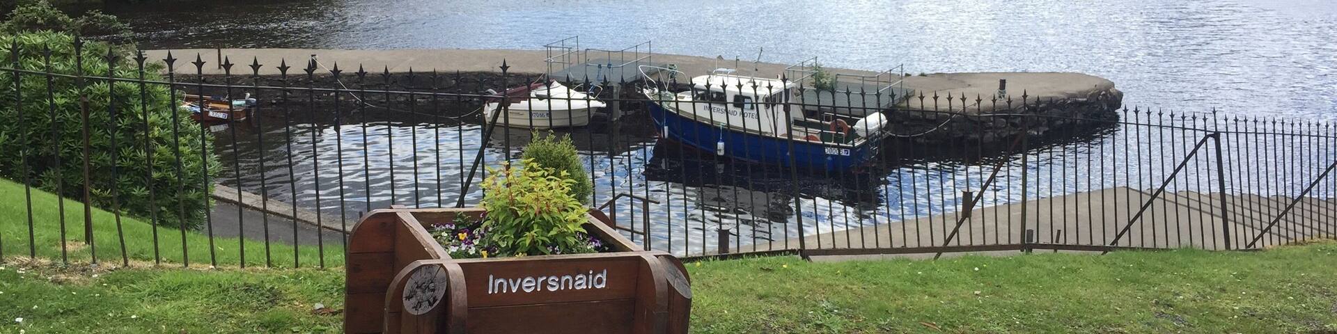 A lake with boats docked, a wooden planter labeled "Inversnaid," and a mountainous landscape in the background.
