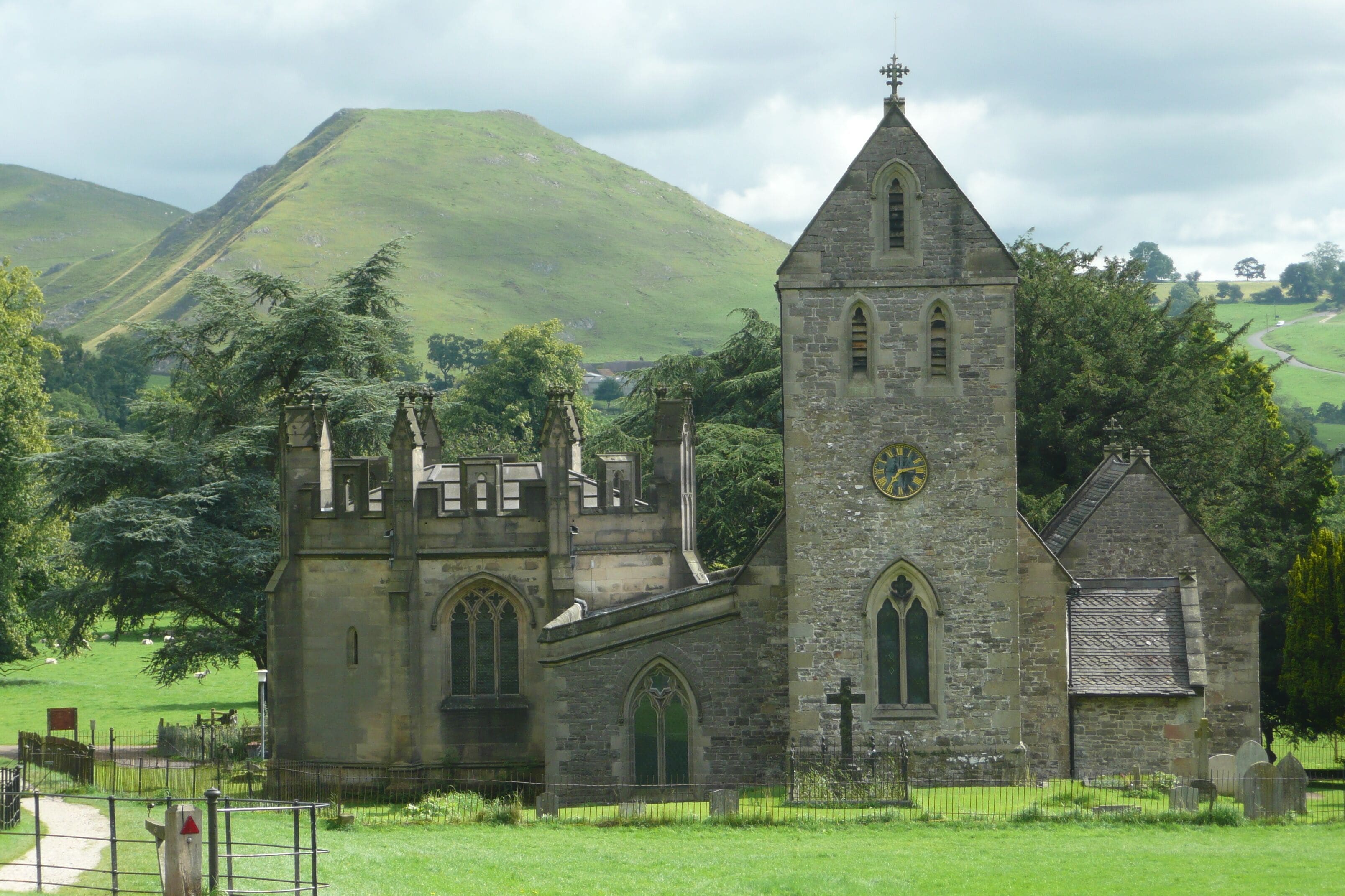 A historic stone church with a clock tower, surrounded by lush green hills and a cemetery.