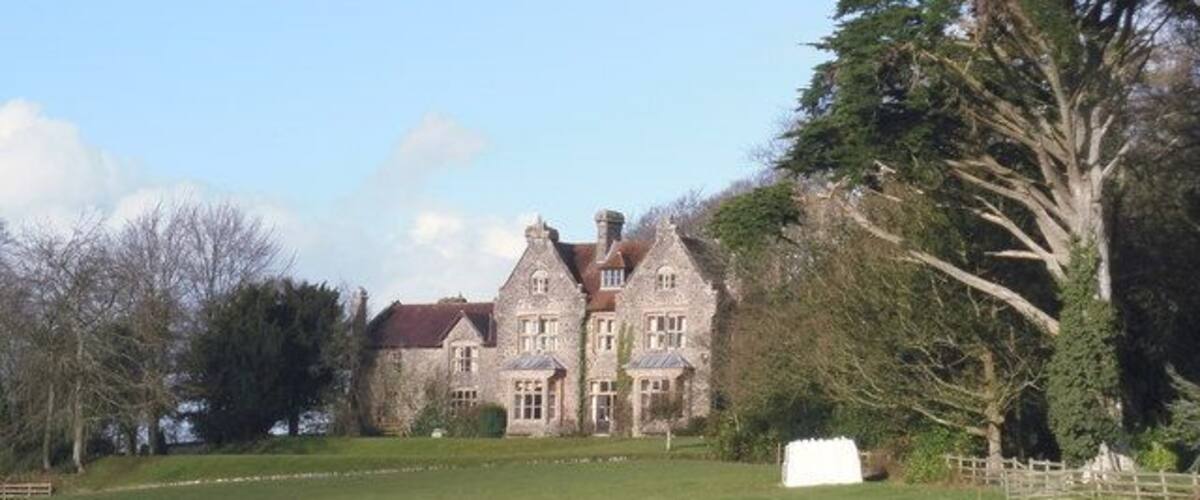 A large, historic stone building with a red roof, surrounded by a well-manicured lawn and trees.