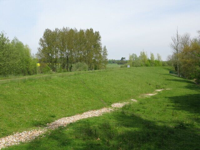 Um campo gramado com um caminho de cascalho, árvores e um edifício ao longe.