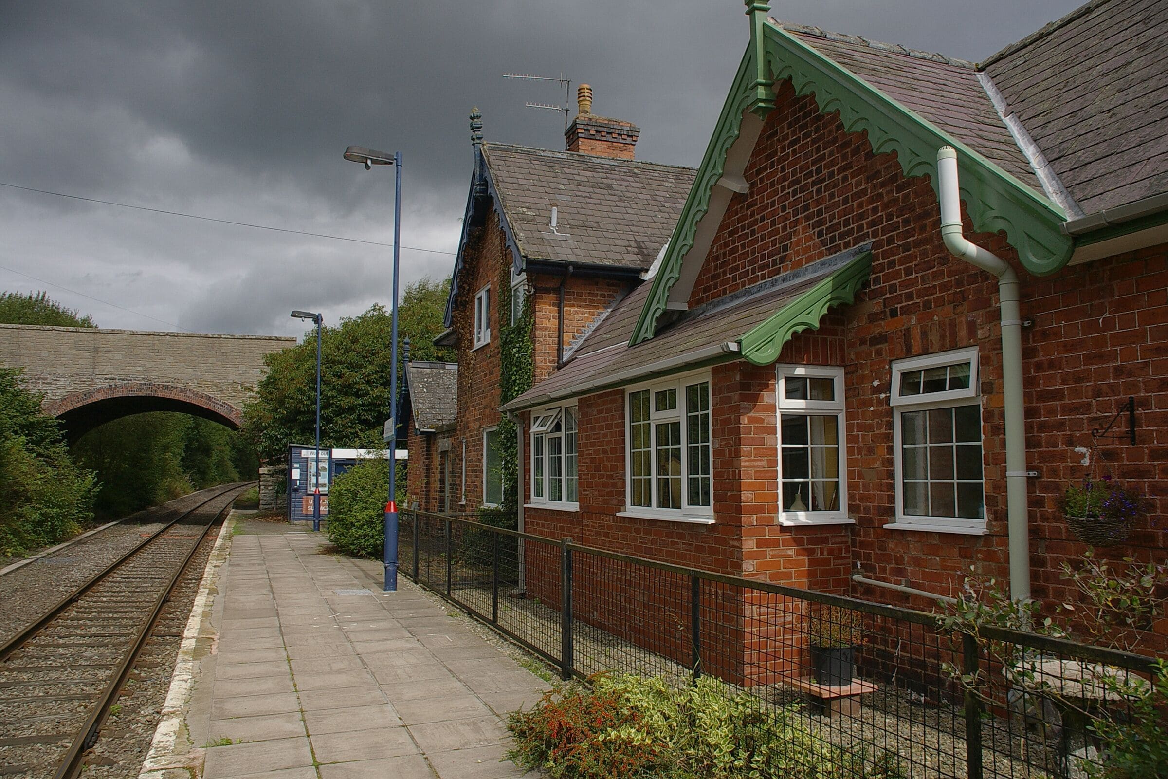 A brick building with a green roof and white windows, a train track, and a stone bridge in the background.