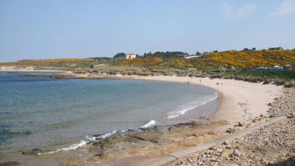 A coastal landscape with a sandy beach, rocky shoreline, and a hill with sparse vegetation.
