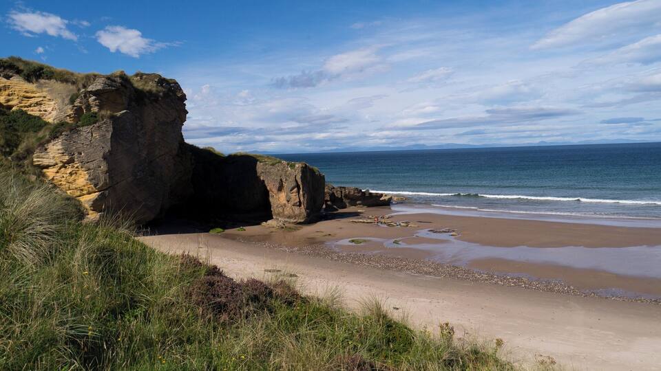 A coastal landscape with a rocky cliff, sandy beach, and ocean waves.