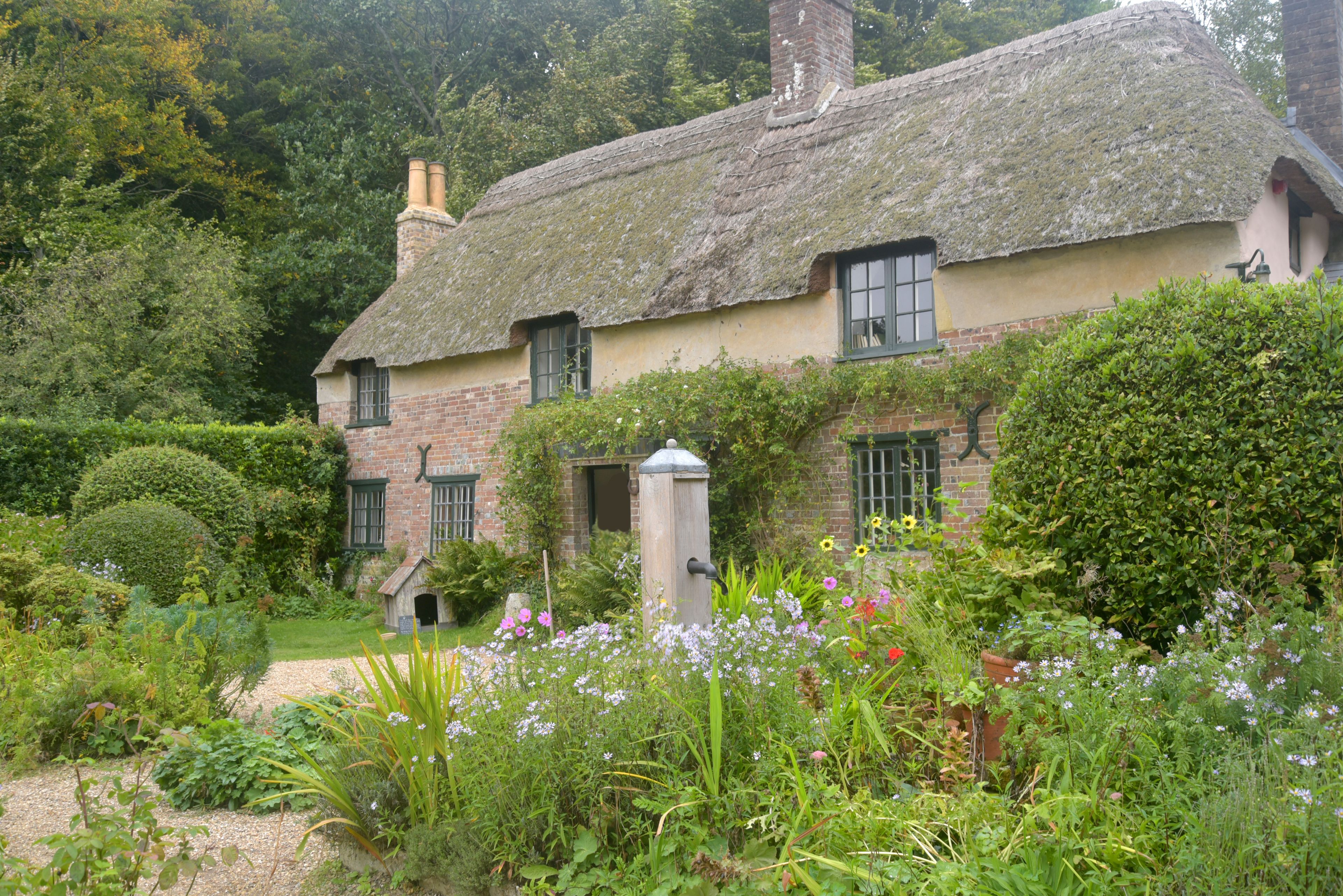 A traditional thatched-roof cottage with a brick chimney, surrounded by a well-maintained garden with various flowering plants and neatly trimmed hedges.