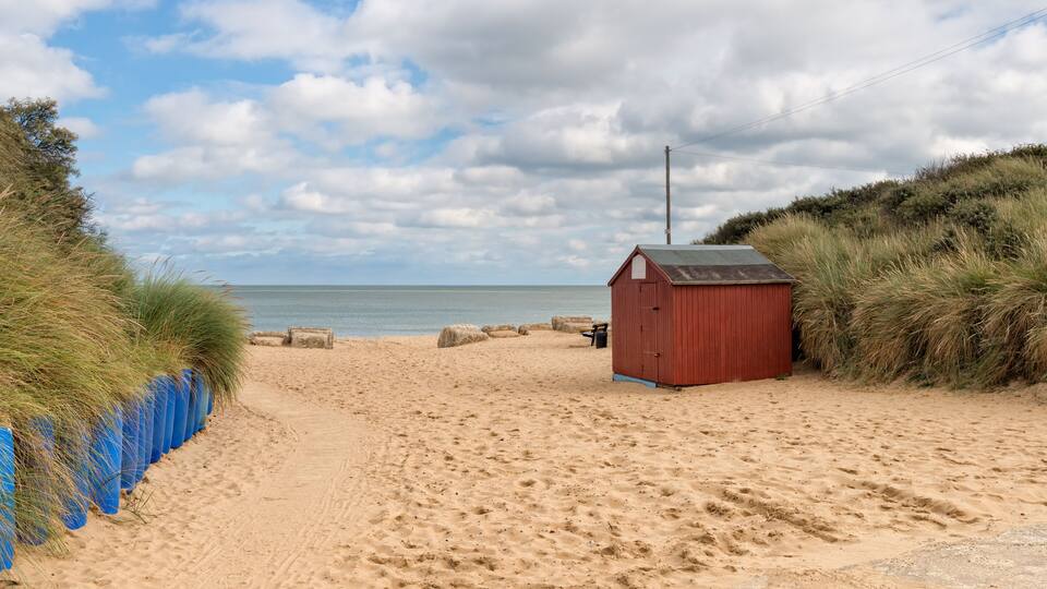 A sandy beach with a red wooden structure, a blue fence, and grass on the dunes.