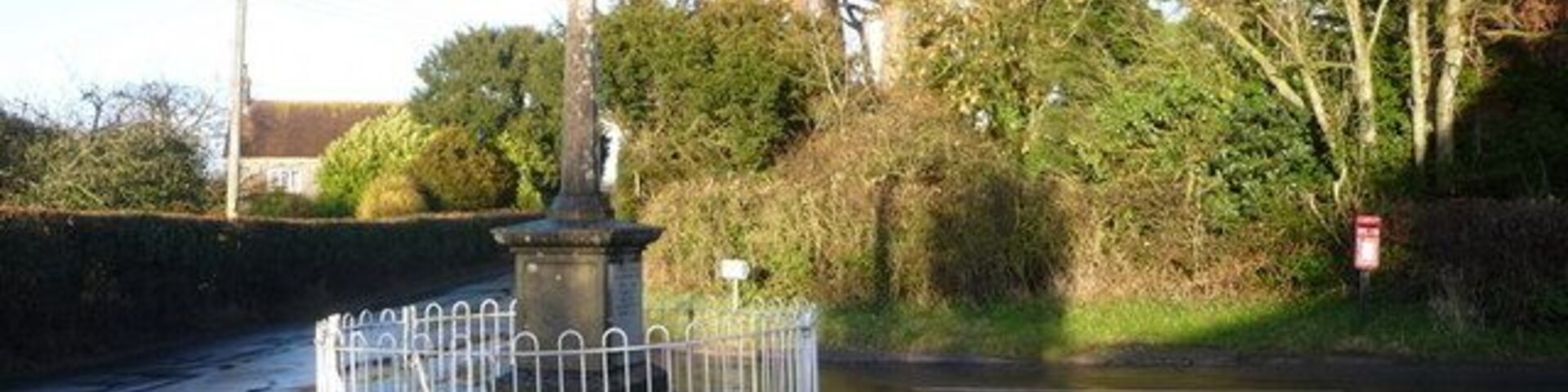 Una rotonda con una isla central, rodeada por una valla metálica, árboles y una carretera con líneas discontinuas blancas.