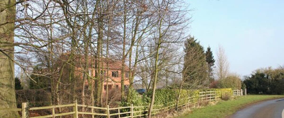 A rural road with a wooden fence, trees, and a house in the background.