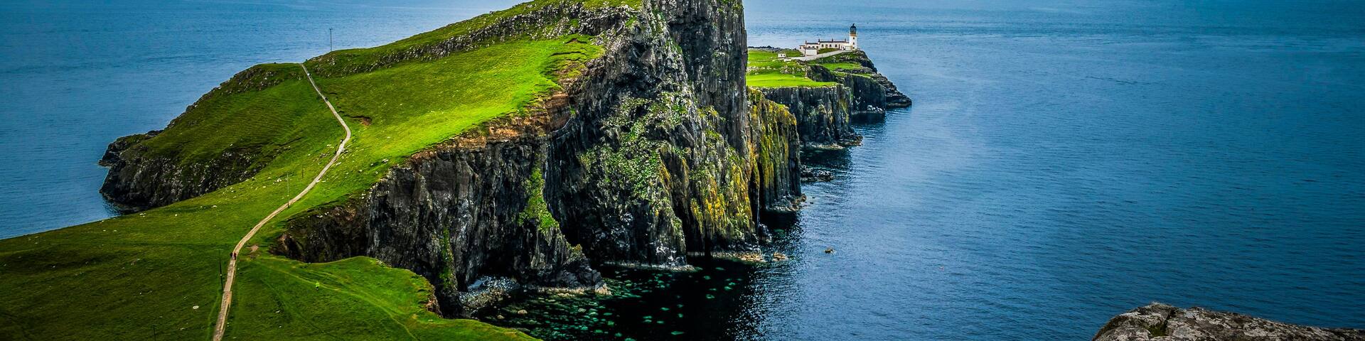 A rugged cliff with a lighthouse on top, surrounded by a clear blue sea and a grassy path leading up to the cliff.