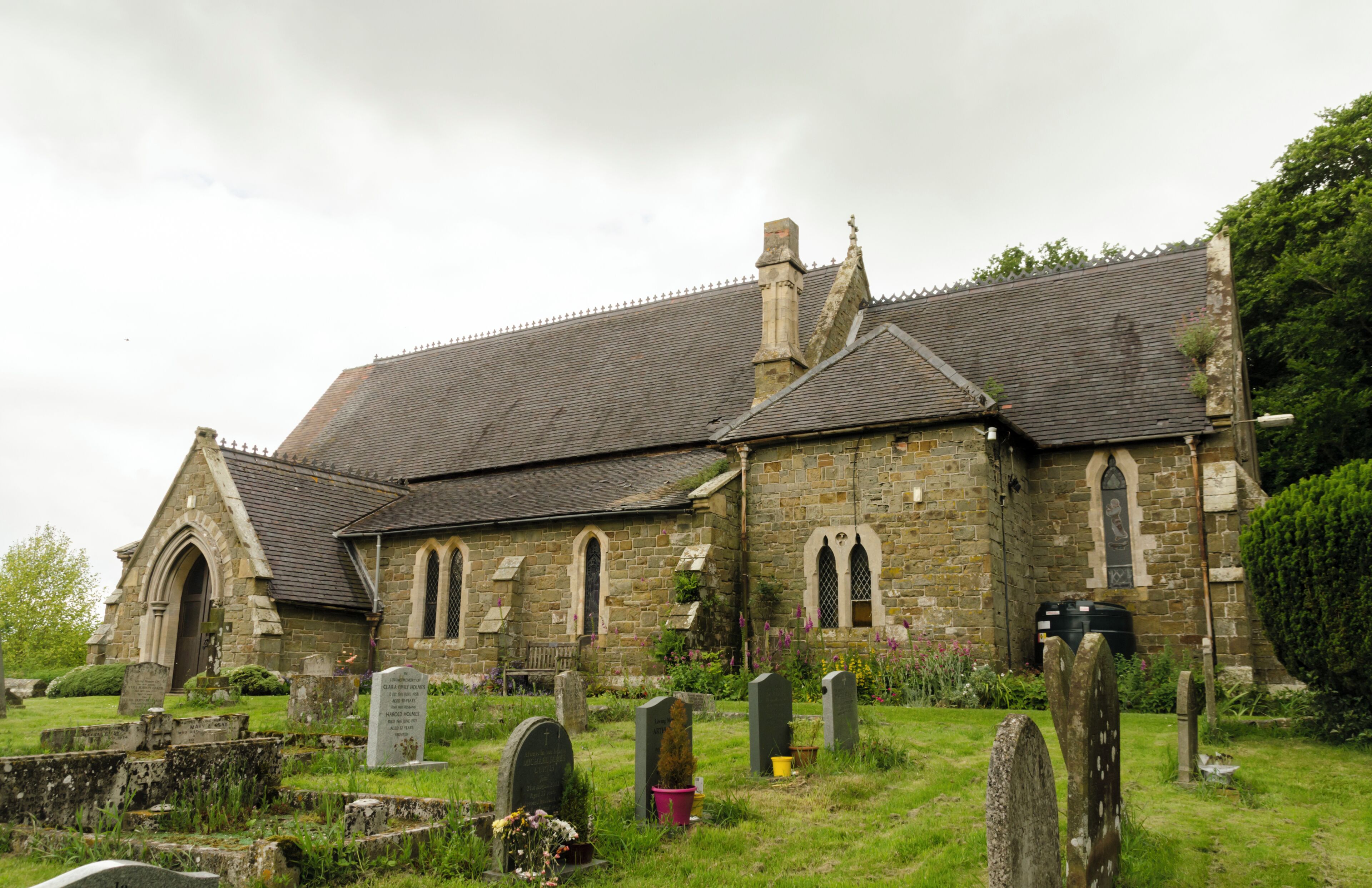 A stone church with a tiled roof, surrounded by a cemetery with headstones and flowers.