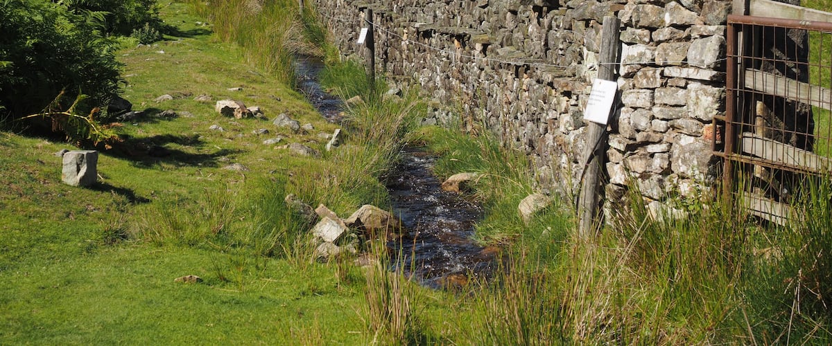 A stone wall with a stream running alongside it, surrounded by lush green hills and a clear sky.