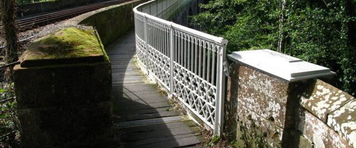 A wooden footbridge with metal railings over railway tracks, surrounded by greenery.
