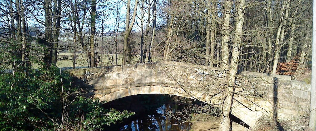 A stone bridge over a narrow waterway, surrounded by trees and bushes.