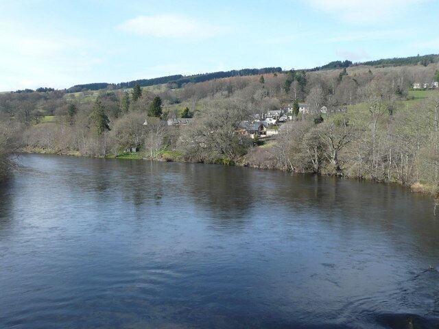 A river with a town on the opposite bank, surrounded by hills and sparse trees.