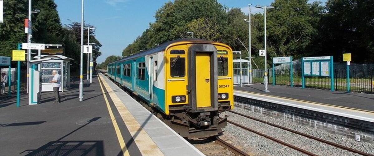 A yellow and blue train at a station platform with trees and a clear sky in the background.