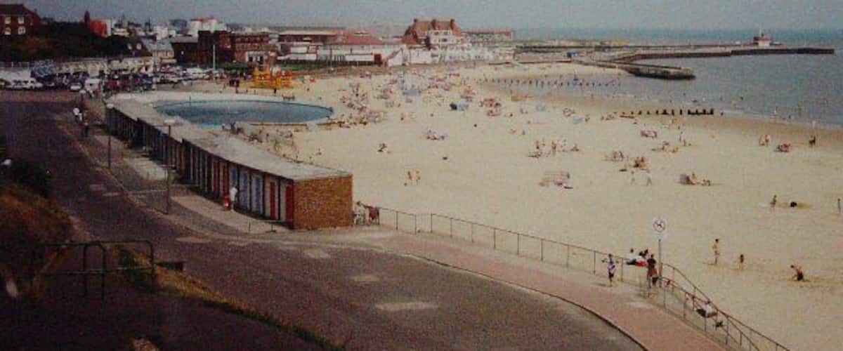 A beach with people, a pool, and buildings in the background.