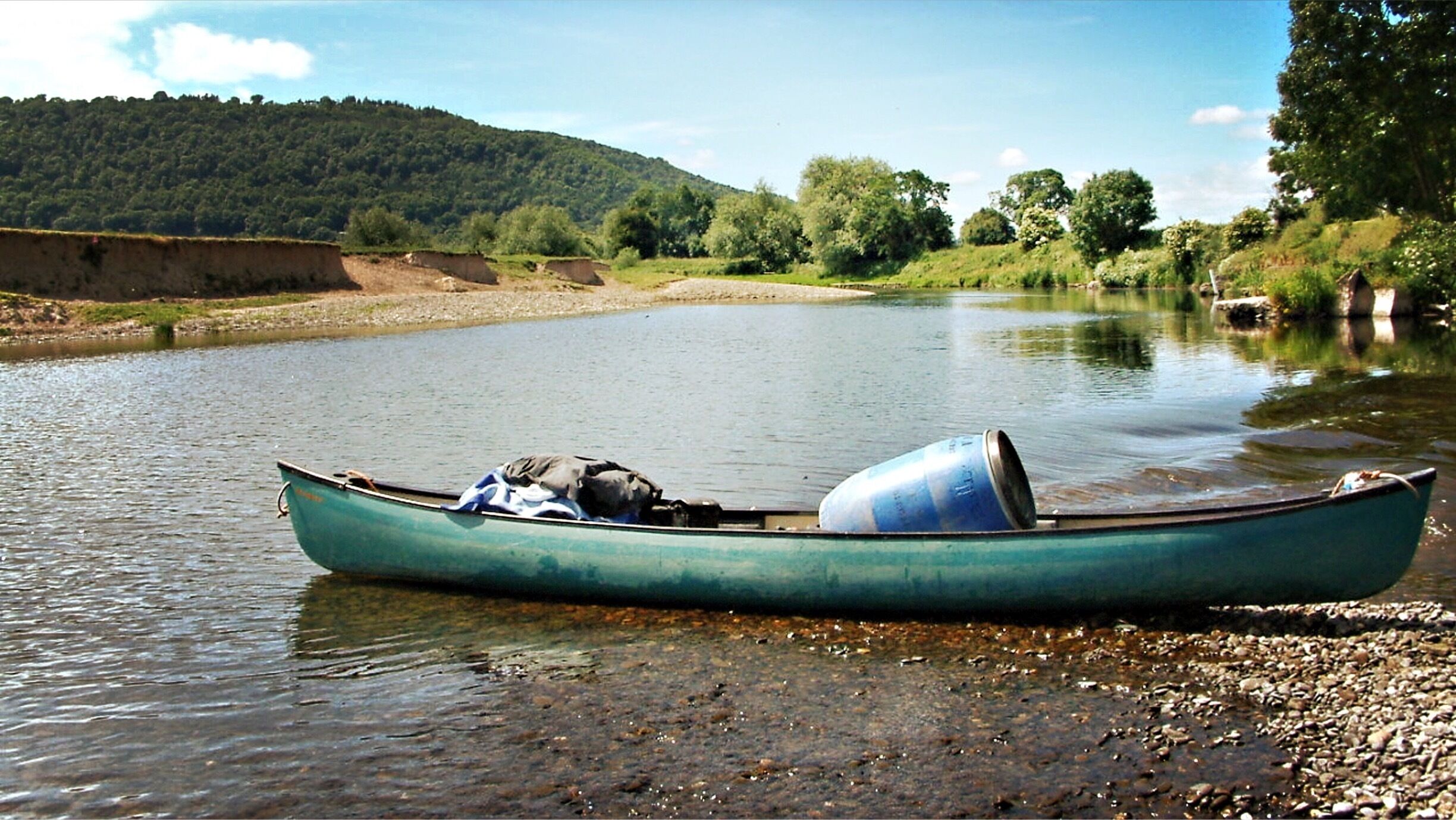 A green canoe on a calm river with a forested hillside in the background.