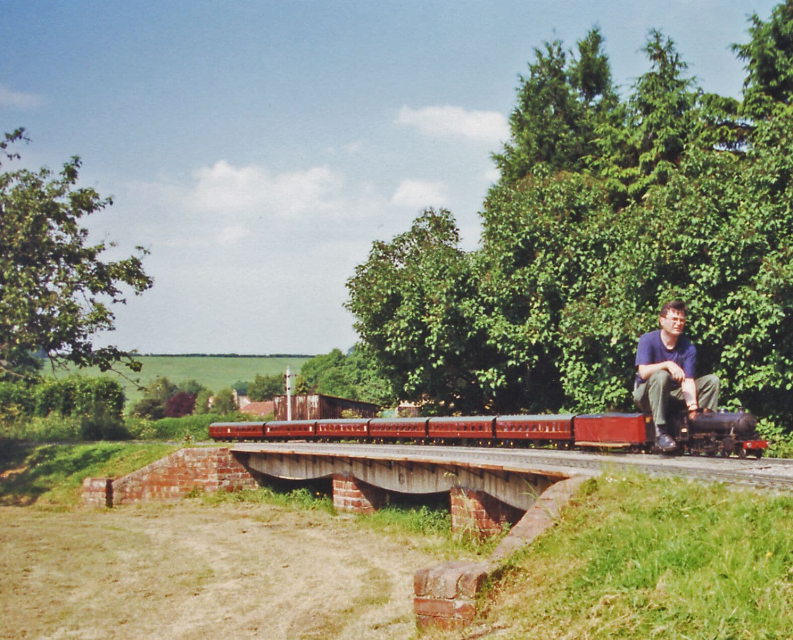 A man sitting on a bridge over a railway track, surrounded by greenery and a clear sky.