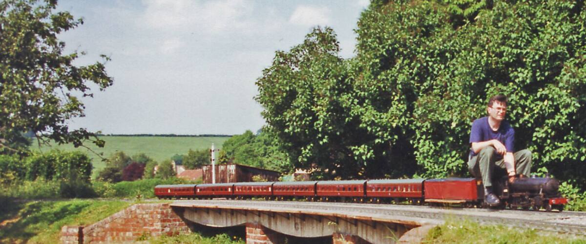 A man sitting on a bridge over a railway track, surrounded by greenery and a clear sky.