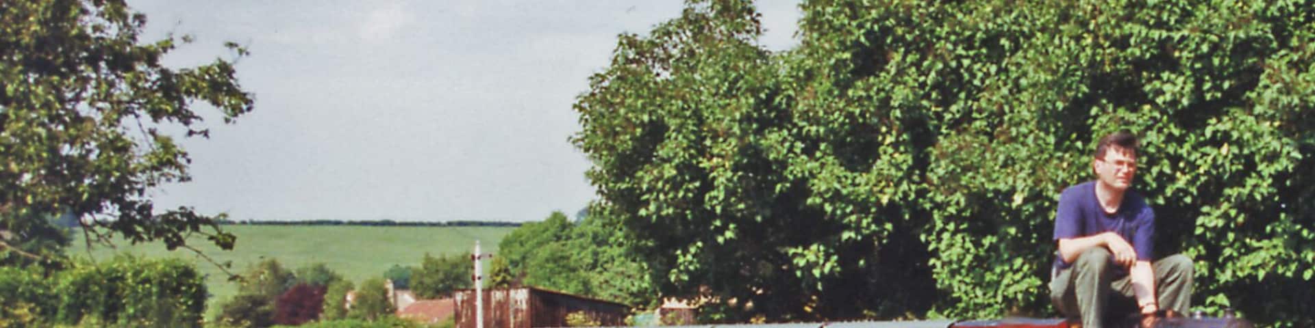 A man sitting on a bridge over a railway track, surrounded by greenery and a clear sky.
