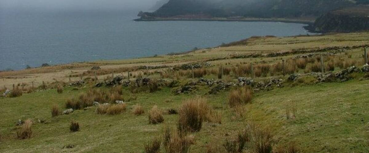 A coastal landscape with a grassy field, a stone wall, and a body of water under a cloudy sky.