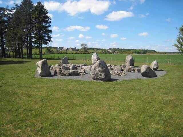 Un círculo de piedras en un campo cubierto de hierba, con árboles y casas al fondo.