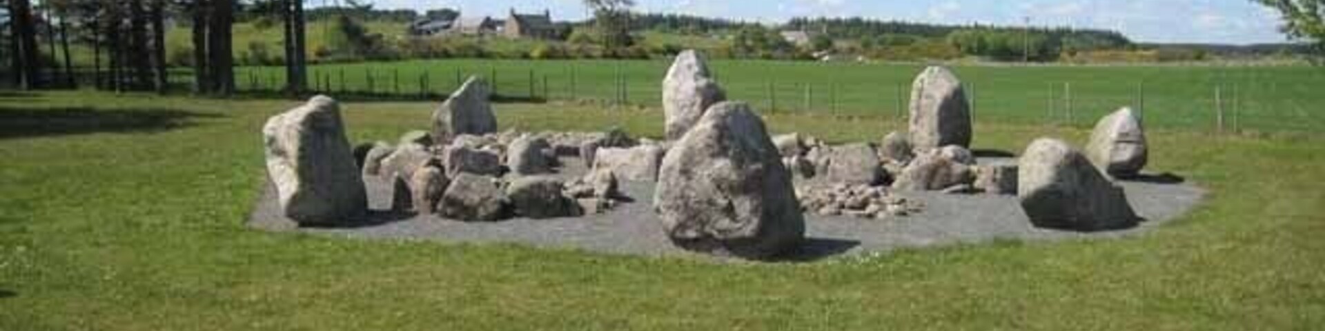 Un círculo de piedras en un campo cubierto de hierba, con árboles y casas al fondo.