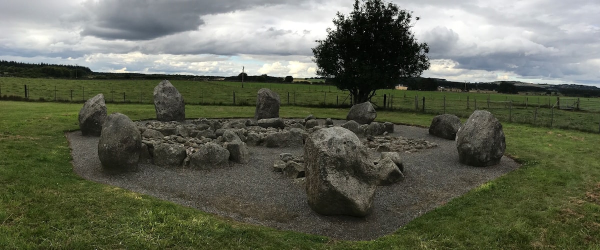 A stone circle with a tree in the center, surrounded by a grassy field and a cloudy sky.