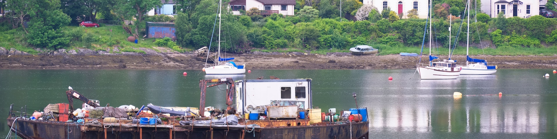 Un bote oxidado amarrado en un puerto tranquilo, con casas y abundante vegetación al fondo.
