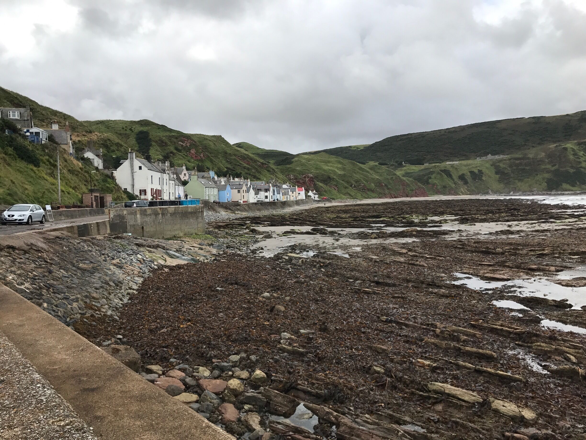A coastal town with houses along the shore, rocky beach, and hills in the background.