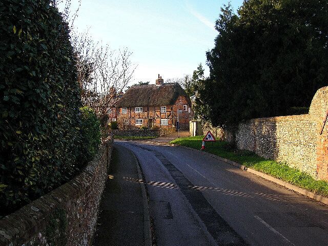 A narrow rural road flanked by stone walls and a thatched-roof house.