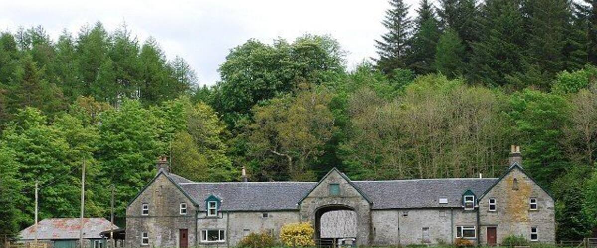 A stone farmhouse with a large arched doorway, surrounded by a lush green field and dense forest.