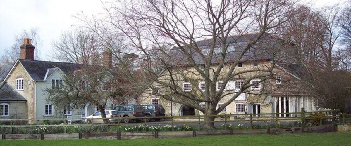 A large estate with a prominent tree in the foreground, a field, and a building with multiple windows and a chimney.