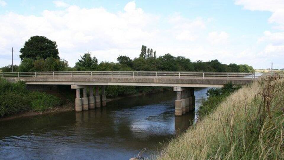 A concrete bridge over a river with grassy banks and trees in the background.