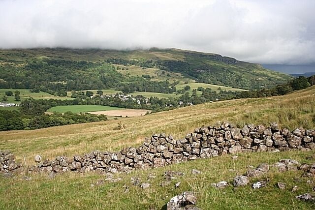 On the Slopes of Dunmore The rickety-looking drystane dyke, marked on the 1:25,000 map, separates the rough grazing from the gully carved by the Kilewnan Burn, behind me. At the other side of the glen the Fintry Hills are partly obscured by low cloud. Fintry itself is among the trees in the middle distance.
