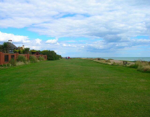 A grassy field with a few people in the distance, a building with a red roof, and a beach in the background.