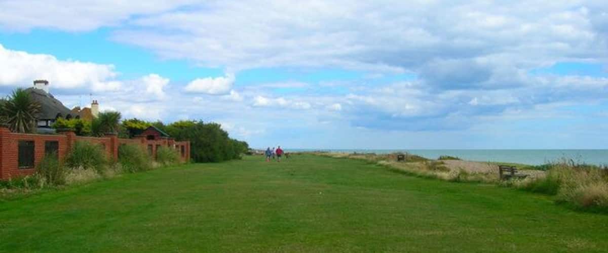 A grassy field with a few people in the distance, a building with a red roof, and a beach in the background.