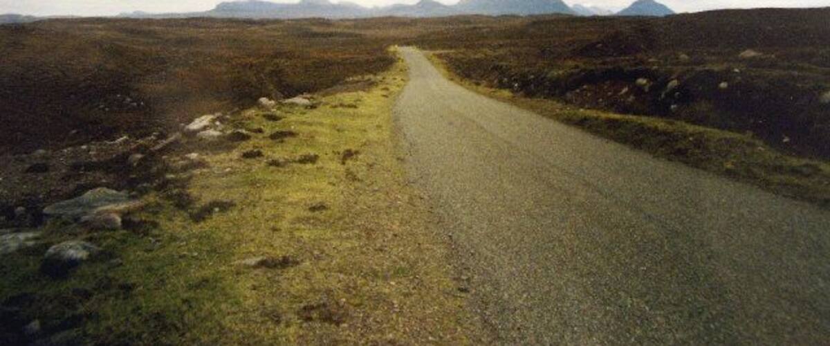 A rural road winding through a barren landscape with distant mountains under a cloudy sky.