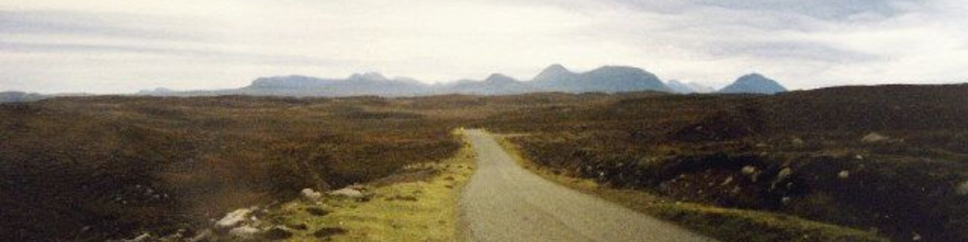 A rural road winding through a barren landscape with distant mountains under a cloudy sky.