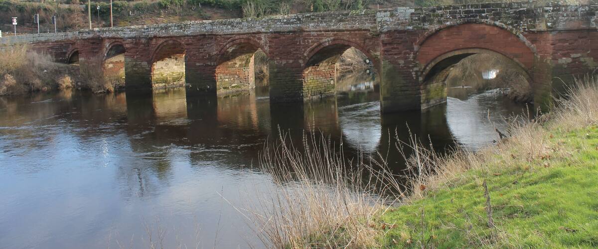 A brick bridge over a calm river with houses in the background.