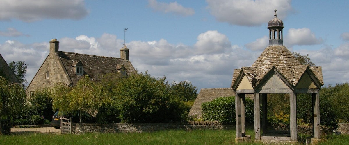 A stone gazebo with a pointed roof and a bell tower in a grassy field.