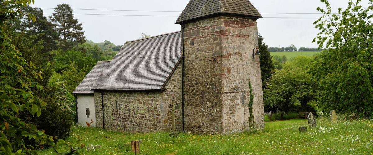 A stone church with a bell tower and a weather vane, surrounded by lush greenery and a grassy field.